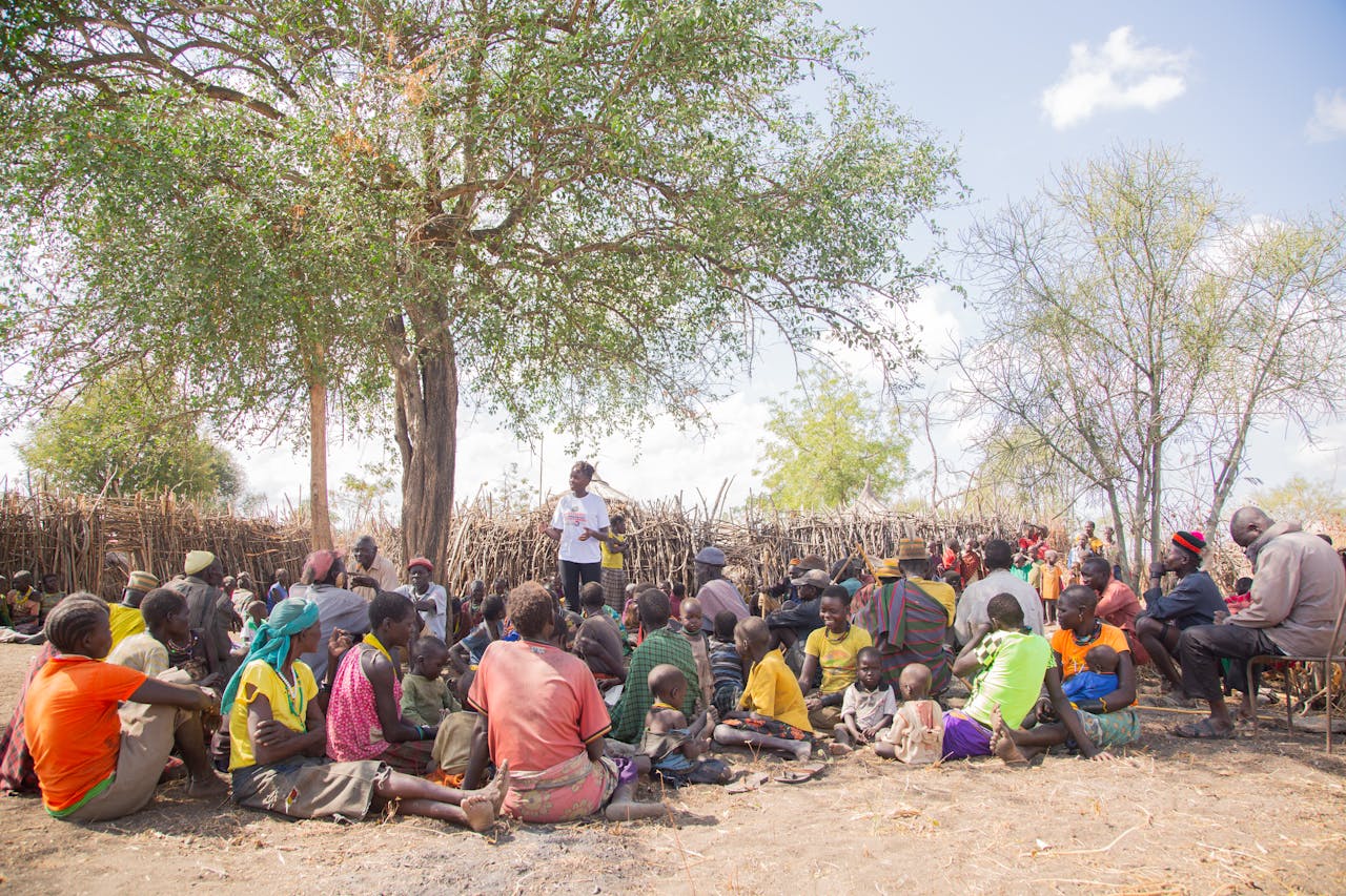 A diverse group of adults and children gather under a tree for a community meeting outdoors.