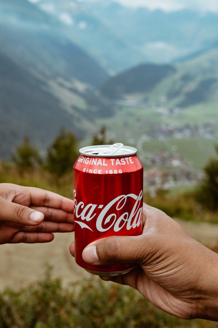 Hands sharing a Coca-Cola can with a scenic mountain backdrop.